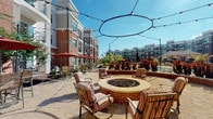 This outdoor lounge is a thoughtfully designed communal gathering space that blends comfort and style within a vibrant courtyard setting. The patio is laid with an elegant stone paver surface throughout, lending a refined, cohesive aesthetic across the entire area.

At the heart of the open-air section sits a circular brick fire pit, surrounded by cushioned lounge chairs with bold striped upholstery — ideal for relaxed conversation or evening gatherings. Overhead, a circular halo of string lights adds an ambient, social atmosphere after dark.

Adjacent to the fire pit area, multiple high-top bistro tables shaded by rich red market umbrellas provide casual dining and socializing options, with direct views of the nearby pool. The poolside setting is framed by lush, colorful plantings that add vibrancy to the space.

A covered pavilion area anchors one side of the lounge, featuring brick-accented columns, wicker seating with plush cushions, and ornamental side tables — offering shade and shelter while remaining open to the surrounding courtyard views. Iron railing borders the pavilion's perimeter, providing a sense of enclosure without obstructing the scenic backdrop of the surrounding multi-story brick buildings. The entire space is bathed in natural light under open skies, creating an inviting, resort-like atmosphere.