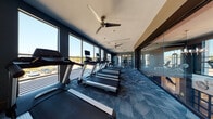 This contemporary fitness center is thoughtfully designed to support a wide range of workout routines in a bright, energizing environment. The main cardio area stretches along a wall of floor-to-ceiling windows, flooding the space with natural light and offering open outdoor views. A row of treadmills and a rowing machine are positioned to take full advantage of this expansive window line, creating an inviting atmosphere for cardio training. Ceiling fans mounted overhead provide consistent airflow throughout the workout floor.

The strength training zone features a multi-functional cable machine, free weights, and additional equipment neatly organized on open shelving units, offering users a comprehensive selection of resistance training options. A dedicated spin studio, separated from the main floor by a full glass wall partition, houses a fleet of stationary cycling bikes and is accented by a bold chevron-patterned floor in blue, brown, and white tones. Wall-to-wall mirrors and a mounted flat-screen television complete this studio space.

Throughout the fitness center, patterned blue carpet tiles in a geometric design add a modern, polished aesthetic that ties the space together. The combination of dark navy accent walls, light neutral ceilings, and ample natural light gives the facility a sleek, professional feel suited for daily use.