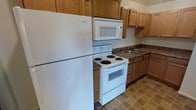 This single-story space includes a well-equipped kitchen that serves as a central feature. The kitchen is outfitted with warm-toned oak cabinetry that provides generous storage both above and below the countertops. White appliances, including a full-size Whirlpool refrigerator, an electric coil range with oven, and an over-the-range microwave, are neatly arranged to maximize efficiency within the layout. The dark laminate countertops offer a contrasting aesthetic against the light cabinetry, and a double-basin sink is conveniently positioned beneath upper cabinets along one wall. The flooring features a neutral tile pattern that complements the overall color palette of the kitchen. The cabinetry extends to the upper walls, making use of vertical space for additional storage. The layout follows an L-shaped configuration, providing a practical workflow between the cooking, prep, and washing areas. Overall, the kitchen presents a clean, functional design suited for everyday use, with a cohesive and understated style throughout.