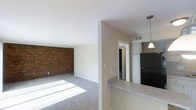This single-floor residence offers a thoughtfully designed open-concept layout that connects the living area and kitchen in a seamless, functional arrangement. The living room is a standout space, anchored by a full exposed brick accent wall that adds texture and warmth to the otherwise neutral, clean aesthetic. Soft carpeting covers the floor throughout the living area, and generous natural light streams in, brightening the space considerably.

A half-wall partition separates the kitchen from the living area while maintaining an open sightline between the two spaces. The kitchen features a laminate countertop breakfast bar, white cabinetry, and a black refrigerator, with a pendant light fixture hanging above to provide focused illumination. Visible HVAC venting indicates climate control throughout the unit.

A hallway leading from the main living area connects to additional rooms, with white paneled doors and neutral beige walls maintaining a consistent, cohesive palette throughout. The combination of the brick wall, soft carpeting, and ample natural light gives the space a comfortable and inviting ambiance suited to everyday living.