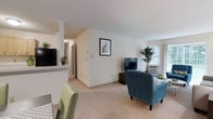 This single-floor residential unit offers a well-organized and functional layout designed for comfortable everyday living. The living area is a central highlight, featuring plush wall-to-wall carpeting that adds warmth underfoot and a neutral color palette of soft whites and beiges that creates a clean, welcoming ambiance throughout. Large windows allow natural light to fill the space, contributing to an airy and open feel.

The living room provides ample space for seating and entertainment, with room for multiple chairs and a media setup. A framed art piece adorns one of the walls, adding a decorative touch to the otherwise understated décor. A window-mounted air conditioning unit ensures year-round comfort.

Adjacent to the living area, the kitchen is partially separated by a low counter or pass-through, maintaining visual connectivity while defining each zone. The kitchen features light-colored countertops and standard appliances visible from the living space.

A hallway leads from the main living area toward additional rooms, including what appear to be bedrooms and a bathroom, offering a clear and intuitive flow through the unit. The corridor is well-lit with overhead lighting. Overall, this unit combines practical design with a bright, neutral aesthetic suited to a variety of lifestyles.