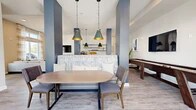 Located at 1880 SW 43rd St in Gainesville, Florida, this single-floor community lounge and kitchen space offers a thoughtfully designed open-concept layout that blends comfort with contemporary style. Striking navy blue structural columns define the space architecturally, creating natural zones within the open floor plan while adding a bold visual contrast against the light-toned walls and wood-look flooring that runs throughout.

The kitchen and bar area features a sleek white countertop island lined with upholstered gray barstools, illuminated by a trio of statement pendant lights with matte black and gold interiors. A herringbone-patterned accent wall serves as a refined backdrop, complemented by open floating shelves and a dedicated coffee and beverage station with white cabinetry.

The living area is anchored by a large sectional sofa, a wall-mounted television, and a linear electric fireplace set against a crisp white feature wall. An elegant gold fringe chandelier adds a touch of glamour overhead. Floor-to-ceiling windows dressed in sheer striped curtains flood the space with natural light and provide views of the surrounding palm-lined exterior. A dining nook with a rounded wood table and upholstered chairs, along with a shuffleboard table, round out the amenities, making this a welcoming and versatile gathering space.