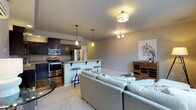 This single-floor residence showcases an open-concept layout that integrates the kitchen, dining, and living areas into one cohesive and functional space. The kitchen is outfitted with dark espresso cabinetry, stainless steel appliances including a French door refrigerator, microwave, and range, and is anchored by a peninsula with a light wood countertop. Industrial-style bar stools provide casual seating along the peninsula, making it ideal for entertaining or everyday dining. Pendant lighting above the peninsula and a flush-mount ceiling fixture illuminate the space warmly. The living area features plush, light-toned sectional seating and is lit by a small chandelier, adding a refined touch. Neutral gray walls and a mix of hardwood and carpeted flooring define the distinct zones while maintaining visual continuity throughout. A rustic wooden cabinet unit along the wall adds warmth and character to the living space. The overall ambiance is modern yet inviting, with a neutral color palette and carefully selected fixtures that balance style and practicality throughout the open floor plan.