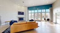 This single-floor community clubhouse offers a well-appointed gathering space designed for both relaxation and social interaction. The centerpiece of the space is a generously sized lounge area distinguished by soaring ceilings and striking teal accent walls. A tiled fireplace with a wall-mounted flat-screen television anchors one end of the room, while a rich tan leather sofa and coordinating blue seating create an inviting conversation area. Wide French doors flood the space with natural light and open directly to an outdoor pool area, seamlessly connecting indoor and outdoor living.

Adjacent to the main lounge, a kitchenette and bar area features light wood-tone flooring, a countertop breakfast bar with seating for two, pendant lighting, and ample counter space — ideal for casual dining or entertaining. A separate dining nook with round tables and upholstered chairs provides additional seating, accented by decorative sunburst mirrors on the wall.

A fully equipped fitness center is accessible directly from the common area, visible through an open doorway and featuring cardio equipment on patterned carpet flooring. Throughout the space, consistent light wood-tone flooring, recessed lighting, and a neutral color palette with bold teal accents create a cohesive, modern aesthetic.