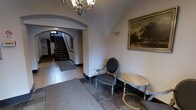 This single-storey space presents a distinguished entryway characterized by its classic architectural details. A prominent arched passageway frames the view toward a staircase beyond, adding depth and a sense of grandeur to the entrance hall. The vaulted ceiling transitions elegantly through the arch, lending the space a timeless, traditional character.

Overhead, a decorative multi-arm chandelier provides warm ambient lighting, complementing the wall-mounted sconce visible beneath the arch. The flooring combines stone tiles and a carpeted runner, offering both visual contrast and practical underfoot comfort. Dark baseboard trim provides a crisp definition between the walls and floor throughout.

A classic upholstered armchair with carved wooden frame and nailhead detailing sits alongside a light wood circular side table, creating an intimate seating vignette. A large framed landscape painting adorns the wall, reinforcing the space's traditional aesthetic. The neutral white walls, arched detailing, and carefully chosen furnishings together establish an atmosphere that is refined, composed, and quietly sophisticated — characteristic of a well-appointed traditional building entrance.