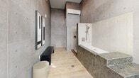 Located in Phoenix, AZ, this thoughtfully designed bathroom delivers a bold, contemporary aesthetic through a carefully curated selection of materials and architectural details. Large-format concrete-look tiles clad the walls from floor to ceiling, lending an industrial yet refined character to the space. Light wood-toned plank flooring provides a warm contrast to the cooler grey tones throughout.

The standout feature is a raised soaking tub platform, finished entirely in a sophisticated herringbone mosaic tile in muted earth tones, accessed by a set of tiled steps that double as a design statement. Adjacent to the tub, a white herringbone-tiled accent wall frames a sleek sliding bar shower fixture. A glass-enclosed shower stall is positioned nearby, offering a separate bathing zone.

A wall-mounted mini-split climate control unit ensures year-round comfort. A floor-to-ceiling window and glass door fill the space with abundant natural light while providing views of the surrounding outdoor environment. Seating is thoughtfully incorporated via a minimalist matte black bench and a cylindrical stool, enhancing the spa-like ambiance. Abstract wall art adds a curated finishing touch, making this bathroom a truly distinctive and functional retreat.