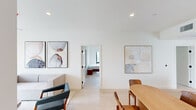 This thoughtfully designed single-story residence offers five bedrooms and three bathrooms across a well-organized floor plan that balances communal living with private retreats. The open-concept kitchen and dining area serves as the heart of the home, anchored by a sleek island with bar seating, stainless steel appliances, and white cabinetry that extends to the ceiling. Light wood flooring runs throughout, reinforcing the space's clean, contemporary aesthetic. An oval dining table with warm wood chairs provides ample seating adjacent to the kitchen, creating a seamless flow for everyday meals and entertaining. The living room features a wall-mounted TV, sliding glass doors that open to a balcony with open views of the surrounding neighborhood, and abundant natural light. Each bedroom maintains the home's minimalist character, with crisp white walls, light-filled windows framing outdoor views, and tile or light wood flooring. The three bathrooms are finished to a high standard, featuring glass-enclosed walk-in showers, light wood vanities with stone countertops, and neutral tile work. Recessed lighting throughout the home enhances its modern, airy ambiance, while curated wall art adds warmth and personality to the shared spaces.