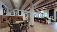 This thoughtfully designed single-floor amenity space combines multiple distinct zones into a cohesive, social environment. The main lounge area is anchored by exposed dark wood ceiling beams that contrast handsomely against white walls and light herringbone-patterned hardwood flooring. A built-in leather banquette with high-top bistro tables creates an intimate gathering nook, accented by a curated gallery wall of vibrant pop-art prints. A dedicated game table with upholstered chairs adds to the relaxed, social atmosphere.

A full wet bar runs along one side of the main area, lined with leather counter stools beneath a row of windows that invite natural light. Adjacent to this, a café station features a built-in coffee machine, dark cabinetry, a large wall-mounted TV, and a striking geometric-tiled floor that distinguishes the space visually.

A separate private dining or meeting room accessed through sliding barn doors offers a substantial solid wood communal table surrounded by wire-frame chairs, illuminated by a cluster of globe pendant lights. A wall-mounted TV and a cozy built-in bench nook with a glass entry door complete this versatile room. Throughout, recessed lighting and carefully selected fixtures reinforce the space's modern yet welcoming character.