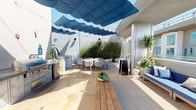 Located at 201 W Lexington Dr in Glendale, CA, this thoughtfully designed rooftop terrace offers an elevated outdoor living experience with stunning panoramic views of the surrounding cityscape. The space is anchored by a bold, scalloped blue shade canopy that stretches overhead, providing ample coverage while adding a distinctive visual character to the environment.

The outdoor kitchen is a central feature, equipped with a stainless steel built-in grill, counter space, and additional appliances, making it well-suited for entertaining. Adjacent to the kitchen, a large dining table surrounded by stylish woven chairs accommodates generous group gatherings, while a wood-accented bench adds warmth and contrast to the contemporary design.

A dedicated lounge area offers a sleek built-in sofa with navy blue cushions and accent pillows, complemented by a tower patio heater for year-round comfort. Lush greenery, including potted tropical plants, sculptural succulents, and a manicured hedge backdrop, softens the modern aesthetic and adds a natural, vibrant element throughout.

A separate open-air balcony extends the space further, featuring high-top tables with succulent centerpieces and far-reaching views of the Glendale skyline under open sky. String lights add an ambient touch, while frosted glass panels and polished column accents reinforce the space's sleek, modern architectural identity.