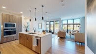 Located at 13125 Watertown Plank Road in Elm Grove, Wisconsin, this thoughtfully designed 2-bedroom, 3-bathroom residence offers a modern aesthetic with high-quality finishes throughout. The open-concept living area seamlessly connects the kitchen, dining room, and living room, all bathed in abundant natural light from expansive floor-to-ceiling windows with sleek black frames that frame outdoor views. The kitchen is a standout feature, boasting two-tone cabinetry with warm wood lower cabinets and crisp white uppers, a gas cooktop, stainless steel wall ovens, a statement range hood, and a large island with pendant lighting — ideal for both cooking and entertaining. The dining area sits adjacent to a glass door leading to the private patio, enhancing the indoor-outdoor flow. The living room features built-in wood shelving and generous wall-to-wall windows. The spacious primary bedroom offers plush carpeting, ample natural light, and a well-appointed en-suite bathroom with a double vanity and a glass-enclosed shower. The second bedroom is bright and comfortable with its own closet. Three full bathrooms, in-unit Whirlpool laundry, and multiple closets complete this refined and functional residence.