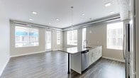 This single-floor space showcases a thoughtfully designed open-concept layout that seamlessly integrates the kitchen and living areas. The kitchen serves as a central focal point, anchored by a generous island with a dark countertop surface and a contrasting bar-height extension supported by sleek black metal legs. White shaker-style cabinetry lines the island, complemented by a stainless steel dishwasher and a modern faucet with an undermount sink. Pendant lighting with exposed Edison-style bulbs hangs above the island, adding an industrial touch.

The flooring throughout features rich, dark hardwood-style planks that contrast elegantly with the neutral gray walls and white trim. Exposed ductwork along the upper walls reinforces the space's contemporary, loft-inspired aesthetic. Recessed lighting is strategically placed across the white ceiling to ensure even illumination throughout.

Large windows dressed with white horizontal blinds line the perimeter, flooding the open living area with natural light. A glass-paned door provides direct access to an outdoor area, enhancing the sense of openness and connectivity to the exterior.