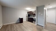 This single-floor unit offers a clean, modern aesthetic throughout, combining functional design with contemporary finishes. The open-concept living and kitchen area is anchored by warm-toned luxury vinyl plank flooring that flows seamlessly across the space, complemented by crisp light gray walls and white baseboards that lend a fresh, updated feel.

The kitchen is a standout feature, outfitted with rich dark espresso cabinetry fitted with sleek brushed nickel hardware. A white countertop provides ample workspace, while a stainless steel single-basin sink with a modern pull-down faucet serves as a practical centerpiece. The kitchen is fully equipped with a stainless steel refrigerator, a freestanding range with oven, and a microwave. A dark gray subway tile backsplash adds a refined, contemporary touch, and an overhead range hood completes the cooking area. A compact dark-toned cabinet unit in the open living zone offers additional storage or surface space.

The bathroom features gray tile flooring and a full tub-shower combination with white tile surround accented by a decorative border stripe, finished with brushed nickel fixtures throughout.

The bedroom is spacious and filled with natural light from a wide double window fitted with blinds, and continues the same vinyl plank flooring. Baseboard heating runs along the perimeter, ensuring comfort year-round.