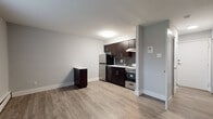 This single-floor unit offers a clean, modern aesthetic throughout, combining functional design with contemporary finishes. The open-concept living and kitchen area is anchored by warm-toned luxury vinyl plank flooring that flows seamlessly across the space, complemented by crisp light gray walls and white baseboards that lend a fresh, updated feel.

The kitchen is a standout feature, outfitted with rich dark espresso cabinetry fitted with sleek brushed nickel hardware. A white countertop provides ample workspace, while a stainless steel single-basin sink with a modern pull-down faucet serves as a practical centerpiece. The kitchen is fully equipped with a stainless steel refrigerator, a freestanding range with oven, and a microwave. A dark gray subway tile backsplash adds a refined, contemporary touch, and an overhead range hood completes the cooking area. A compact dark-toned cabinet unit in the open living zone offers additional storage or surface space.

The bathroom features gray tile flooring and a full tub-shower combination with white tile surround accented by a decorative border stripe, finished with brushed nickel fixtures throughout.

The bedroom is spacious and filled with natural light from a wide double window fitted with blinds, and continues the same vinyl plank flooring. Baseboard heating runs along the perimeter, ensuring comfort year-round.