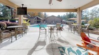 Located at 1811 Sibley Rd in Augusta, Georgia, this single-level covered outdoor pavilion serves as a welcoming communal gathering space designed for relaxation and socializing. The structure features a classic brick exterior with sturdy support columns and a white-painted ceiling equipped with ceiling fans to keep the space comfortable year-round. A standout focal point is the full brick wood-burning fireplace, complete with a rustic wooden mantel and a large decorative clock above it, flanked by Adirondack chairs arranged on a vibrant botanical-patterned area rug — creating a cozy and inviting lounge atmosphere. Multiple bar-height tables and chairs are distributed throughout the pavilion, offering versatile seating options for casual dining or conversation. Mounted flat-screen televisions enhance the entertainment experience. The pavilion opens directly to a resort-style outdoor pool area, offering expansive views of the shimmering water, lounge chairs, and umbrella-shaded tables set against a lush backdrop of mature trees, manicured hedges, and colorful seasonal plantings. The seamless transition between the covered pavilion and the open pool deck makes this space ideal for outdoor gatherings in any season.