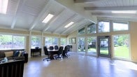 This single-floor space makes a strong architectural impression with its striking vaulted ceiling defined by exposed white-painted beams that rise to a dramatic peak. Skylights and flush fluorescent fixtures run along the ceiling's ridge, ensuring the interior is consistently well-lit throughout the day. The open floor plan is unified by light-toned tile flooring that stretches across the entire space, lending a clean and polished feel underfoot.

Expansive windows wrap around the perimeter, inviting generous amounts of natural light and offering unobstructed views of the lush, tree-lined outdoor surroundings. A glass-paneled entry door further connects the interior to the verdant exterior, enhancing the sense of openness and accessibility.

The space is organized into distinct functional zones. A comfortable lounge area features plush seating arranged for relaxed conversation, complemented by framed artwork on the warm yellow-toned walls. A dedicated conference area includes a substantial meeting table surrounded by ergonomic office chairs, making it well-suited for professional gatherings or collaborative sessions. A compact dining or breakout area with a smaller table and chairs rounds out the layout.

Overall, the space conveys an airy, welcoming ambiance that balances professional utility with casual comfort, making it highly adaptable for a variety of uses.