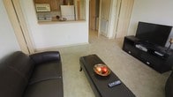 This single-floor residence offers a well-designed, open-concept layout that seamlessly connects the main living area to the kitchen. The living room is carpeted and comfortably sized, providing ample space for seating and entertainment, with a media stand positioned conveniently along one wall. A pass-through counter with a light wood finish bridges the living area and the kitchen, making the space feel open and connected while maintaining distinct functional zones. The kitchen is equipped with standard appliances, including a white refrigerator and microwave, set beneath warm-toned wooden cabinetry. Neutral-colored walls and light carpeting throughout contribute to a bright, clean ambiance. A hallway visible from the main living area leads further into the unit, hinting at additional rooms beyond. Interior doors with classic paneled detailing add a traditional architectural touch, while a visible HVAC vent reflects practical utility. Overall, the space presents a straightforward, functional residential layout with a neutral palette that offers a versatile canvas for personalization.
