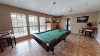 Located at 7771 Joe Newton St in San Antonio, Texas, this single-floor game room is purpose-built for recreation and entertainment. The centerpiece of the space is a full-size pool table with green felt, finished in sleek black and accented by a stylish three-shade pendant light that hangs directly overhead, ensuring excellent illumination during play. A dedicated wall-mounted cue rack keeps an assortment of billiard cues neatly organized and within easy reach.

The room is finished with warm terracotta-toned tile flooring throughout, lending the space a casual yet polished aesthetic. Neutral, taupe-painted walls are complemented by vertical wood-panel detailing on select walls, adding a touch of texture and character. Recessed lighting throughout the ceiling supplements natural light, which streams in through multiple windows fitted with horizontal blinds.

A compact high-top bistro table with a granite-style surface and upholstered stools offers a comfortable spot for spectators or casual conversation. Framed artwork decorates the walls, contributing to the room's relaxed, lounge-like ambiance. Windows along one side of the room provide direct views of an outdoor swimming pool, seamlessly connecting indoor entertainment with outdoor leisure. A doorway leads to an adjoining interior space, making this room an accessible and central hub within the home.