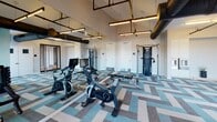 The Vestry Bloomfield Ave Fitness Center in Montclair, NJ is a thoughtfully designed workout facility that combines industrial aesthetics with modern functionality. The space is defined by its bold herringbone-patterned carpet tiles in shades of teal, gray, and white, which anchor the room with a vibrant, contemporary feel throughout.

Exposed ductwork in matte black runs along the ceiling, complementing the suspended linear pendant lighting that illuminates the space efficiently. High ceilings contribute to an open, airy atmosphere, while a teal accent wall adds a dynamic visual focal point. Full-wall mirrors on one side amplify the sense of space and are a practical addition for form-checking during workouts.

The fitness center is well-equipped with Peloton spin bikes, elliptical machines, a cable/functional trainer system, a rowing machine, a full dumbbell rack, and a weight bench, offering a comprehensive range of cardio and strength training options. A wall-mounted television provides entertainment during workouts.

Multiple large windows allow abundant natural light to filter in and offer views of the surrounding Montclair area. A glass-paneled door provides access to an adjacent outdoor terrace. The entrance area includes built-in storage and a mirrored entry, creating a clean and welcoming transition into the facility.