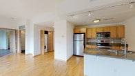 This single-floor residence is designed with a clean, modern aesthetic throughout. The open-concept living area is anchored by light hardwood flooring and large floor-to-ceiling sliding glass doors that lead to a private balcony, flooding the space with natural light and providing views of the surrounding neighborhood's greenery and streetscape. An additional window further enhances the brightness of the living area.

The kitchen is well-equipped with light maple cabinetry, granite countertops, a subway tile backsplash, and a full suite of stainless steel appliances, including a refrigerator, electric range, and built-in microwave. A peninsula counter provides additional workspace and casual seating potential, while the kitchen flows seamlessly into the main living area.

A hallway connects to the bedroom, which features plush wall-to-wall carpeting, a window with an outdoor view, and access to a spacious walk-in closet with built-in shelving. In-unit laundry, housed behind louvered doors, adds everyday convenience.

The bathroom is finished with dark and light tile flooring in a checkered pattern, a granite-topped vanity with maple cabinetry, a large mirror, and a tub-shower combination with sliding glass doors. Track lighting and recessed fixtures provide consistent illumination throughout the unit.