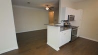 This single-story space showcases an open-concept design that seamlessly connects the kitchen and living areas. The kitchen is equipped with stainless steel appliances, including a gas range and over-the-range microwave, complemented by white shaker-style cabinetry and dark gray countertops. A peninsula island with matching cabinetry and countertop extends into the space, providing additional prep area and serving as a natural divider between the kitchen and the adjoining living area. A tiled backsplash adds a decorative accent behind the range. Dark hardwood flooring runs consistently throughout both the kitchen and living area, creating a cohesive and grounded aesthetic. Crown molding lines the upper walls, adding an architectural detail that elevates the overall finish of the space. The living area is generously sized and features a ceiling fan with a light fixture, contributing to both air circulation and ambient lighting. Neutral-toned walls keep the space feeling bright and versatile, while a paneled entry door with sidelights is visible in the background, hinting at the entryway beyond.