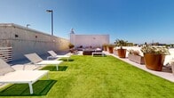 Located at 1700 Newbury Park Dr in San Jose, CA, this rooftop terrace offers an expansive open-air retreat designed for both relaxation and entertaining. The space is anchored by a generous stretch of vibrant artificial turf, providing a verdant centerpiece that contrasts beautifully with the clean, light-toned concrete deck surrounding it. A dedicated lounge area features a large sectional sofa and accent chairs with plush cushions, arranged to maximize comfort and social interaction. Sun loungers are positioned along the turf for those looking to enjoy the abundant California sunshine. The dining zone is outfitted with a substantial slatted wood table and woven rope chairs, ideal for al fresco meals, and is situated adjacent to a full-size gas BBQ grill for outdoor cooking. Numerous large planters—ranging from warm copper-toned vessels to sleek concrete troughs—are distributed throughout the terrace, filled with ornamental trees, grasses, and shrubs that add texture, color, and a sense of privacy. The rooftop enjoys panoramic views of the surrounding San Jose cityscape and the rolling Diablo Range mountains in the distance, all under open skies. The overall design aesthetic is modern and resort-inspired, combining natural elements with contemporary furnishings to create a sophisticated and inviting outdoor environment.