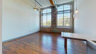 This single-floor space showcases a compelling fusion of industrial heritage and contemporary design. The open-concept layout is anchored by soaring ceilings that emphasize the generous volume of the interior, while original hardwood floors with rich, warm tones run throughout, lending a sense of continuity across the space.

The main living area is a standout, featuring dramatic arched windows framed by exposed brick walls that flood the room with natural light and offer views of surrounding greenery. An exposed metal ductwork system and wooden ceiling beams add to the industrial aesthetic, complemented by a ceiling fan and track lighting overhead.

The kitchen is well-appointed with gray shaker-style cabinetry, marble-look quartz countertops, and a full suite of stainless steel appliances including a French door refrigerator, electric range, and over-the-range microwave. A kitchen island provides additional workspace and casual seating.

The space includes multiple rooms accessible through paneled gray doors. A notable bedroom features soaring ceilings, a rustic wooden column, and access to a walk-in closet. Two bathrooms offer gray vanities with quartz countertops—one with vessel sink and hardwood flooring, the other with stone-look tile and a round mirror. An interior staircase and a pass-through window add architectural interest throughout.