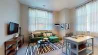 This thoughtfully designed single-floor residence offers a clean, contemporary aesthetic defined by light wood-style flooring, neutral wall tones, and an abundance of natural light throughout. The open-concept living area is anchored by large windows dressed with sheer curtains, allowing soft, diffused light to fill the room and creating an inviting atmosphere ideal for both relaxing and entertaining. The kitchen is well-appointed with two-tone cabinetry featuring soft blue-grey upper cabinets and white lower cabinets, white quartz countertops, a tile backsplash, and a full suite of black appliances including a refrigerator, range, microwave, and dishwasher. A dining area sits adjacent, providing a seamless flow between cooking and dining spaces. The bedrooms are generously proportioned, each featuring large windows that frame views of the surrounding greenery and provide ample natural light. The unit includes two bathrooms, both finished with grey cabinetry, white countertops, and tile flooring. One bathroom features a glass-enclosed walk-in shower with white subway tile, while the other includes a combination layout with a large vanity mirror. The overall design of the space is modern and understated, emphasizing clean lines, functional layouts, and a cohesive neutral palette throughout.