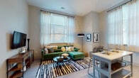This thoughtfully designed single-floor residence offers a clean, contemporary aesthetic defined by light wood-style flooring, neutral wall tones, and an abundance of natural light throughout. The open-concept living area is anchored by large windows dressed with sheer curtains, allowing soft, diffused light to fill the room and creating an inviting atmosphere ideal for both relaxing and entertaining. The kitchen is well-appointed with two-tone cabinetry featuring soft blue-grey upper cabinets and white lower cabinets, white quartz countertops, a tile backsplash, and a full suite of black appliances including a refrigerator, range, microwave, and dishwasher. A dining area sits adjacent, providing a seamless flow between cooking and dining spaces. The bedrooms are generously proportioned, each featuring large windows that frame views of the surrounding greenery and provide ample natural light. The unit includes two bathrooms, both finished with grey cabinetry, white countertops, and tile flooring. One bathroom features a glass-enclosed walk-in shower with white subway tile, while the other includes a combination layout with a large vanity mirror. The overall design of the space is modern and understated, emphasizing clean lines, functional layouts, and a cohesive neutral palette throughout.