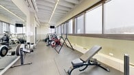 Located at 1975 Boulevard de Maisonneuve Ouest in Montréal, this fitness center offers a well-organized, single-floor exercise facility designed to maximize functionality within a compact footprint. The room follows a long, narrow layout that makes efficient use of the available space, with equipment arranged along both sides to allow for comfortable circulation throughout.

The ceiling features exposed white-painted structural beams with linear LED lighting running along the length of the room, providing consistent, bright illumination. A continuous row of large, horizontally oriented windows lines one full wall, flooding the space with natural light and offering open views of the surrounding Montréal cityscape — a notable feature that enhances the workout environment.

Full-length mirrors on one wall visually expand the room and support proper form during exercise. The flooring consists of durable rubber fitness mats, providing traction and impact absorption. Equipment on-site includes stationary bikes, elliptical trainers, treadmills, a multi-function cable/weight machine, a rowing machine, a weight bench, and a stability ball. A wall-mounted television is also present for entertainment during workouts.

A dedicated exit door is clearly marked ("SORTIE"), ensuring straightforward access and safety compliance. Overall, this fitness center provides a bright, practical, and well-equipped environment suited for a comprehensive individual workout routine.