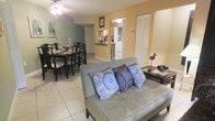 This single-story space is designed with an open-concept layout that connects the living, dining, and kitchen areas into one cohesive and functional environment. The living area features a cushioned loveseat accented with decorative patterned pillows, positioned to face the rest of the shared space, and is complemented by a side table with decorative accents. Light-toned ceramic tile flooring runs consistently throughout, contributing to a clean, airy aesthetic.

Adjacent to the living area, the dining space accommodates a full dining set with a dark-finished rectangular table and matching chairs, ideal for hosting meals. A ceiling fan overhead serves both the dining and living zones, providing practical air circulation. The warm beige and sage-toned walls create a welcoming atmosphere across the open floor plan.

A glimpse into the kitchen reveals a raised countertop bar, light cabinetry, and tile flooring that continues from the main living area, maintaining visual continuity. A set of louvered bi-fold closet doors adds functional storage along one wall. The overall layout and finishes reflect a practical, comfortable design suited for everyday living.