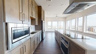 This thoughtfully designed single-floor residence combines contemporary style with high-end functionality. The centerpiece of the home is a striking galley-style kitchen, lined on both sides with custom light wood cabinetry extending to the ceiling for abundant storage. Premium stainless steel appliances — including a built-in microwave, refrigerator, double wall oven, and a cooktop beneath a large stainless steel range hood — sit alongside light quartz countertops and a stainless steel sink. Rich dark hardwood flooring runs the length of the kitchen, which opens toward a bright, window-lined living area with sweeping city views and a ceiling fan overhead. The bedrooms feature plush neutral carpeting and large windows that flood each room with natural light while offering elevated views of the surrounding urban landscape. The primary bathroom is well-appointed with a double-sink vanity featuring white quartz countertops, sleek gray cabinetry, a glass-enclosed shower with floor-to-ceiling tile, and warm wood-look flooring. A hallway connects multiple rooms and additional closet space, reinforcing the unit's practical layout. Throughout the residence, clean lines, neutral tones, and quality materials create a polished, modern ambiance.