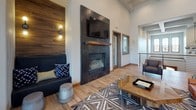 Located at 1100 E Plum Creek Pkwy in Castle Rock, Colorado, this single-floor community clubhouse delivers a thoughtfully designed gathering space that blends rustic warmth with modern sophistication. The open-concept layout is anchored by a spacious lounge area featuring wood-look tile flooring, a striking dark-tiled fireplace with a wood mantel, and a wall-mounted television. An accent wall clad in reclaimed-style wood paneling adds texture and warmth to this inviting seating area. Adjacent to the lounge, a dedicated dining and kitchenette area showcases blue-gray cabinetry, quartz countertops with a subtle veining pattern, a mounted television, an under-counter refrigerator, a wine rack, and open shelving with decorative accents. Large garage-style windows flood this section of the space with natural light and offer views of the outdoors. A cozy reading nook features a dramatic curved window arrangement extending from floor to soaring ceiling height, framing abundant natural light and outdoor views — an ideal spot for quiet retreat. Exposed beam detailing and high ceilings throughout lend the space an airy, grand quality, while the warm wood flooring and carefully chosen furnishings create a welcoming, residential ambiance suited for community gatherings and social connection.