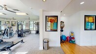 Located at 1400 Village Blvd in West Palm Beach, Florida, this fitness center offers a comprehensive workout facility across a single floor. The space is thoughtfully divided into distinct functional zones, providing a well-rounded exercise experience for users of all fitness levels.

The main cardio and strength area features a row of treadmills and additional cardio machines, including a rowing machine, positioned along large windows that offer views of the outdoor pool and lush Florida landscaping. Natural light fills the room, creating an energizing atmosphere. Multiple ceiling fans ensure comfortable airflow throughout. A multi-station cable machine, a full rack of dumbbells, and various benches provide ample options for strength training. A wall-mounted flat-screen TV allows users to stay entertained during workouts.

A dedicated studio area with hardwood flooring, a ballet barre, and floor-to-ceiling mirrors offers space for stretching, balance training, and group classes. This section is further equipped with stability balls, kettlebells, and medicine balls for functional fitness training. Colorful framed artwork adorns the white walls throughout, adding a vibrant, modern aesthetic. The overall design is clean, bright, and well-organized, making this fitness center a functional and inviting space for daily exercise.