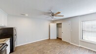 Located at 9524 E 71st St in Tulsa, Oklahoma, this single-floor unit offers a thoughtfully arranged layout comprising a living area and a full bathroom. The main living space is finished with warm wood-look laminate flooring that runs throughout, complemented by neutral gray-toned walls and clean white baseboards that create a bright, cohesive aesthetic. A ceiling fan with a light fixture is centrally mounted beneath a textured ceiling, providing both air circulation and ambient lighting, while recessed lighting adds additional illumination. A built-in kitchenette along one wall features white upper cabinetry, a stainless steel electric range, a refrigerator, and a sink with granite-style countertops — offering convenient cooking essentials in a compact footprint. A reach-in closet with shelving provides practical storage. A window with blinds allows natural light to fill the room. The bathroom is well-equipped with a large vanity topped with granite-style countertops, a wide mirror with overhead lighting, white cabinetry, and a walk-in shower stall fitted with grab bars for added accessibility. Tile flooring completes the bathroom's clean, functional design. Overall, the space presents a practical and comfortable environment suited to efficient, everyday living.