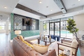 Located in Miami, FL, this thoughtfully designed single-floor community clubhouse offers a generous, open-concept layout that seamlessly blends social, dining, and recreational spaces. Wood-look tile flooring flows throughout, grounding the space with a warm, contemporary aesthetic.

The lounge area is a standout, featuring exposed rustic wood ceiling beams that contrast beautifully against crisp white walls. A tufted leather sofa, spindle-back armchairs, and vibrant colorful rugs create an inviting atmosphere, while a large-format art piece and decorative wall niches add curated character. An intricate laser-cut screen frames a wall-mounted TV above gleaming teal mosaic tile, adding a striking focal point.

The dining area accommodates a large communal table beneath ornate brass pendant lights, perfect for group gatherings. Nearby, a bar-height counter with leather stools lines the floor-to-ceiling black-framed windows, offering direct views of the outdoor pool. Natural light pours in abundantly throughout the day, creating a bright and airy atmosphere.

A green mosaic tile accent column anchors the kitchen/bar area, which includes cabinetry and beverage storage. The space also features a billiards table, rounding out the recreational offerings. With direct glass door access to the pool area, this clubhouse delivers a seamlessly connected indoor-outdoor living experience.