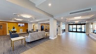 This expansive single-floor community space is thoughtfully designed to foster social engagement and comfort. The main entrance features large glass doors that flood the tiled foyer with natural light, creating a bright and welcoming arrival experience. A staircase and elevator provide convenient access to upper levels, framed by textured wall coverings and clean architectural lines.

The open layout flows seamlessly from the entry into multiple distinct lounge and gathering areas, each appointed with comfortable upholstered seating arrangements, patterned carpeting, and warm accent walls in rich golden yellow and soft grey tones. Recessed lighting and large drum-style ceiling fixtures provide consistent, inviting illumination throughout.

A dedicated game room features a full-size billiards table, dining-height tables with stylish chairs, and bold accent walls adorned with framed artwork — ideal for recreation and casual socializing. A separate lounge area includes a wall-mounted television, a fireplace, and built-in shelving, offering a cozy space for relaxation or group gatherings.

The overall ambiance is modern yet approachable, combining neutral tones with vibrant accents to create an environment that is both functional and aesthetically refined. This well-appointed community space is clearly designed to support an active and connected lifestyle.