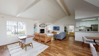 Located at 300 Hilary Way in Vallejo, CA, this single-story space offers a bright and airy atmosphere defined by dramatic vaulted ceilings with exposed beams and an open-concept layout. Rich hardwood flooring flows throughout, creating a warm and cohesive aesthetic across all areas.

The main living area is expansive and welcoming, anchored by a fireplace and illuminated by track lighting along the ceiling beams. A dedicated dining area sits comfortably alongside, making the space well-suited for both relaxed gatherings and more formal occasions. Large windows invite generous natural light throughout the day.

A secondary lounge area features soaring cathedral ceilings with tall clerestory windows that flood the room with light. Striking red-framed French doors open directly to an outdoor patio, creating a seamless indoor-outdoor connection ideal for entertaining or relaxation.

The galley-style kitchen is clean and functional, featuring white cabinetry, light countertops, stainless steel appliances including a refrigerator, range, and dishwasher, and ample storage. Additional rooms visible from the main living space include a fitness area and a private office or workspace, offering versatility for a range of uses. The overall design balances modern mid-century aesthetics with practical, well-organized functionality throughout.