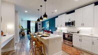 This single-floor residence showcases a bold, eclectic design aesthetic that seamlessly blends vibrant color with modern finishes throughout. The open-concept kitchen is a standout feature, offering crisp white shaker cabinetry, quartz countertops, a textured tile backsplash, and a full suite of stainless steel appliances including a French door refrigerator, range, built-in microwave, and dishwasher. A generously sized central island with seating provides both additional workspace and a casual dining option, illuminated by a trio of dramatic matte black pendant lights. The kitchen flows directly into the living area, which is anchored by a deep teal accent wall and wood-look plank flooring that runs throughout the main living spaces. The living room offers abundant natural light through large windows dressed in floor-length navy drapes and features recessed and chandelier lighting overhead. The bedroom continues the bold color palette with deep gray walls, plush carpet flooring, and large windows that bring in natural light and outdoor views. The full bathroom is equipped with a dual-sink vanity with white cabinetry, dark tile flooring, a soaking tub, and convenient in-unit washer and dryer. Recessed lighting and matte black fixtures complete the refined, contemporary finish of the space.