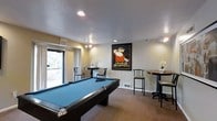 This single-floor recreational space is thoughtfully designed to serve as a social hub for leisure and entertainment. The centerpiece of the room is a full-sized pool table with a striking blue felt surface, providing ample space for gameplay and inviting friendly competition. Surrounding the pool table, the room is furnished with several high-top bistro tables paired with cushioned bar-height chairs, creating comfortable spots for conversation and relaxation. A large window along one wall allows natural light to filter into the space, brightening the neutral-toned walls and textured ceiling fitted with recessed and track lighting. Decorative touches elevate the ambiance, most notably a bold vintage-style framed poster — a "La Bon Bock Atlantique" advertisement — displayed prominently on the wall, adding a classic, café-inspired character to the room. Small floral arrangements on the bistro tables contribute a refined finishing touch. The carpeted flooring adds warmth underfoot, while the overall layout is open and well-organized, ensuring easy movement throughout the space. The combination of functional amenities and tasteful décor makes this a welcoming and versatile gathering area.