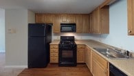 This 2-bedroom, 1-bathroom residence offers a practical and comfortable single-floor layout. The kitchen is a standout feature, equipped with warm honey-toned cabinetry, granite-look countertops, and a full suite of black appliances including a gas range, over-the-range microwave, refrigerator, and dishwasher. Rich hardwood-style flooring grounds the kitchen space, adding warmth and easy maintenance. The open living area transitions seamlessly toward the bedrooms, featuring soft wall-to-wall carpeting and light, neutral wall tones that create a calm and inviting ambiance. Sliding glass doors in the main living area open to a private balcony, allowing natural light to pour in and offering pleasant outdoor views of surrounding greenery. Both bedrooms are generously sized, each with carpeted floors, closet storage, and windows that bring in natural light. Wall-unit air conditioners and baseboard heating ensure year-round comfort. The bathroom is clean and functional, featuring a tiled tub-shower combination, wood-look flooring, a white vanity with sink, and a large mirror. A textured ceiling runs throughout the residence, adding a classic residential character to the space overall.