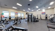 This single-floor fitness facility offers a spacious, open layout designed to accommodate a wide variety of workout routines. The room features a drop ceiling with recessed panel lighting that provides bright, even illumination throughout the space, complemented by natural light streaming in through a row of large windows along one wall, offering views to the exterior.

The flooring consists of durable rubber gym matting, ideal for both heavy equipment and high-impact activities. The facility is outfitted with an impressive selection of equipment, including a central multi-functional cable machine, a squat rack, adjustable weight benches, a full dumbbell rack, a stationary bike, and a variety of plate-loaded machines. Colorful bumper plates add a practical visual element to the strength training area.

Multiple wall-mounted flat-screen TVs are positioned around the room, keeping users entertained during workouts. The neutral, light-colored walls contribute to an open and energizing atmosphere. The overall layout is thoughtfully organized, allowing for comfortable movement between stations and making this facility well-suited for both individual training and group use.