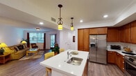 This single-story unit at 2615 Cannon Rd in Carlsbad offers a well-designed open-concept layout with cohesive finishes throughout. The kitchen is a functional centerpiece, featuring warm-toned wood cabinetry extending to the ceiling, stainless steel appliances including a side-by-side refrigerator with ice dispenser, an electric range, and a microwave. A central island with a light quartz countertop and an undermount sink provides additional prep space and is illuminated by a pair of decorative pendant lights with ornate bronze fixtures. Crown molding adds a refined touch.

The open layout connects the kitchen to a dedicated dining area with hardwood-style flooring and a pendant light overhead, and to a living room featuring sliding glass doors and windows that bring in natural light and access to a private outdoor patio. The bedroom continues the connection to the outdoors through its own set of sliding glass doors, opening to a furnished patio. A walk-in closet provides ample storage. The bathroom includes a walk-in shower with grab bars, a wood-toned vanity, and matching wall cabinetry. A separate den or office space with hardwood-style floors offers additional flexibility.