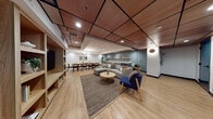 Located on Wilshire Boulevard in the heart of Los Angeles, this single-floor amenity space combines thoughtful design with versatile functionality. The interior features warm light wood flooring throughout, complemented by a distinctive drop ceiling with wood-paneled tile accents and recessed lighting that creates a welcoming, well-lit atmosphere.

The space is divided into several distinct zones. A generous lounge area anchors the main room, furnished with a modular gray sectional sofa, a navy blue accent armchair with wooden legs, and a round coffee table atop a woven area rug. Built-in shelving with light wood cabinetry houses a large wall-mounted television, providing a media-ready gathering spot. A quieter seating nook nearby offers two upholstered accent chairs beside a teal-painted door, ideal for smaller conversations.

The dining area features a long natural wood table accommodating numerous guests, surrounded by black rattan-back chairs and accented by a globe chandelier overhead. Adjacent to the dining space, a full kitchenette is outfitted with gray cabinetry, stainless steel appliances, a beverage refrigerator, and floating wood shelves displayed against a bold teal accent wall. Lush indoor plants are positioned throughout, adding a fresh, organic element to the contemporary Scandinavian-inspired aesthetic.