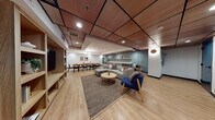 Located on Wilshire Boulevard in the heart of Los Angeles, this single-floor amenity space combines thoughtful design with versatile functionality. The interior features warm light wood flooring throughout, complemented by a distinctive drop ceiling with wood-paneled tile accents and recessed lighting that creates a welcoming, well-lit atmosphere.

The space is divided into several distinct zones. A generous lounge area anchors the main room, furnished with a modular gray sectional sofa, a navy blue accent armchair with wooden legs, and a round coffee table atop a woven area rug. Built-in shelving with light wood cabinetry houses a large wall-mounted television, providing a media-ready gathering spot. A quieter seating nook nearby offers two upholstered accent chairs beside a teal-painted door, ideal for smaller conversations.

The dining area features a long natural wood table accommodating numerous guests, surrounded by black rattan-back chairs and accented by a globe chandelier overhead. Adjacent to the dining space, a full kitchenette is outfitted with gray cabinetry, stainless steel appliances, a beverage refrigerator, and floating wood shelves displayed against a bold teal accent wall. Lush indoor plants are positioned throughout, adding a fresh, organic element to the contemporary Scandinavian-inspired aesthetic.