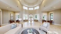 Located at 11411 Colbert Creek Loop in Raleigh, NC, this generously sized single-floor community facility offers a thoughtfully designed layout encompassing multiple functional spaces. The grand foyer serves as the centerpiece of the facility, boasting soaring ceilings, an octagonal architectural form, and floor-to-ceiling sheer-draped windows that flood the space with natural light. Elegant neutral tones, large-format tile flooring, and a central round accent table create a welcoming and sophisticated first impression. Adjacent living areas feature warm hardwood flooring, a tray ceiling, a classic fireplace, and open sight lines to the kitchen bar area. French doors lead to an outdoor patio, further enhancing the sense of openness. A secondary living room with steel-blue walls and plush carpeting provides a quieter, more intimate gathering space with abundant natural light from multiple windows. The dining room continues the blue-toned palette, featuring a round dark wood table surrounded by upholstered chairs and a gallery wall of framed artwork. The kitchen area is equipped with granite countertops, white cabinetry, stainless steel appliances, and a French door opening to the outdoor terrace. The well-equipped exercise room features rubberized flooring, cardio machines, strength equipment, and large windows overlooking the surrounding greenery.