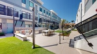Located at 160 S Ventura Ave in Ventura, California, this contemporary residential complex showcases a clean, modern architectural aesthetic defined by a light grey and blue exterior facade, large energy-efficient windows, and private balconies with sleek metal railings. The building's multi-story design is complemented by well-maintained communal outdoor spaces that serve as the heart of the community.

The central courtyard is a standout feature, offering a spacious outdoor gathering area complete with a built-in barbecue station, an outdoor dining table with ample seating, and a comfortable lounge area with cushioned bench seating — ideal for entertaining or relaxing under the Southern California sun. Lush turf lawns, drought-tolerant plantings, and wood-chip garden beds are thoughtfully integrated throughout the grounds, providing both visual appeal and low-maintenance greenery.

Concrete walkways connect various areas of the complex, including side passages with carefully landscaped borders featuring native shrubs and ornamental grasses. Stairways with sturdy metal handrails provide access to upper levels, and string lights draped across the courtyard add a warm ambiance during evening hours. The overall design balances modern urban living with inviting communal spaces, all situated in the vibrant coastal city of Ventura.