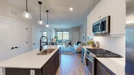 This thoughtfully designed single-floor residence showcases a seamless open-concept layout that connects the kitchen and living area, creating an inviting and functional living space. The kitchen is a standout feature, boasting a two-tone cabinet design with crisp white upper cabinets and bold navy lower cabinets, complemented by a large island with a white quartz countertop and an undermount sink. Stainless steel appliances, including a gas range, microwave, and French door refrigerator, add a professional touch, while globe pendant lights overhead contribute a modern aesthetic. Light wood-style flooring runs throughout the main living areas, enhancing the bright and airy atmosphere. The living area flows naturally from the kitchen and is filled with natural light from large windows and sliding glass doors that open to an outdoor space. The bedroom is a comfortable retreat, carpeted for warmth, with multiple windows offering views of the surrounding area. A dedicated walk-in closet with wire shelving provides ample storage. Neutral tones and clean lines are carried throughout the space, creating a cohesive, contemporary feel that is both practical and visually appealing.