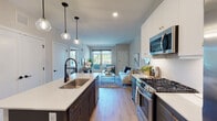 This thoughtfully designed single-floor residence showcases a seamless open-concept layout that connects the kitchen and living area, creating an inviting and functional living space. The kitchen is a standout feature, boasting a two-tone cabinet design with crisp white upper cabinets and bold navy lower cabinets, complemented by a large island with a white quartz countertop and an undermount sink. Stainless steel appliances, including a gas range, microwave, and French door refrigerator, add a professional touch, while globe pendant lights overhead contribute a modern aesthetic. Light wood-style flooring runs throughout the main living areas, enhancing the bright and airy atmosphere. The living area flows naturally from the kitchen and is filled with natural light from large windows and sliding glass doors that open to an outdoor space. The bedroom is a comfortable retreat, carpeted for warmth, with multiple windows offering views of the surrounding area. A dedicated walk-in closet with wire shelving provides ample storage. Neutral tones and clean lines are carried throughout the space, creating a cohesive, contemporary feel that is both practical and visually appealing.