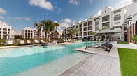 This outdoor amenity space is designed to deliver a resort-like experience, centered around a generously sized swimming pool with clear, inviting turquoise water. The pool area is framed by a wide, tiled sun deck lined with sleek lounge chairs and shade umbrellas, providing ample space for relaxation. Towering palm trees are strategically placed throughout, adding a tropical aesthetic that enhances the overall ambiance. A shallow wading area with mosaic tile accents and colorful planters with flowering accents adds a refined touch to the poolside environment. Adjacent to the pool, a stretch of well-maintained turf provides a clean, green contrast to the hard-surface deck. A dedicated outdoor patio area offers multiple round dining tables with woven chairs, strung with overhead bistro lighting and set against a strikingly textured stone and large-format glass facade. The surrounding multi-story buildings feature a modern architectural style with white and gray exteriors, stepped gabled rooflines, and private balconies that overlook the courtyard. The entire space is enclosed by decorative wrought-iron fencing, creating a defined and secure environment while maintaining an open, airy feel under the open sky.