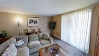 This single-floor space presents a cozy and functional living room as its centerpiece. The room is finished with wall-to-wall carpeting in a neutral gray tone, contributing to a warm and understated aesthetic throughout. Walls are painted in a soft beige, creating a calm, neutral backdrop that complements the overall design.

A large sliding glass door dressed with full-length vertical blinds dominates one wall, allowing for generous natural light while offering privacy control. The blinds provide a clean, classic look and suggest access to an outdoor area beyond.

The living room is furnished with a light sage-green sofa set arranged around a wood-and-glass coffee table, creating an inviting seating arrangement suited for relaxation or casual gathering. A console table along one wall displays decorative accents, including vases and a framed artwork depicting an architectural scene, adding a touch of character to the space.

A tall potted tree plant positioned in the corner introduces a natural element, enhancing the room's warmth. A baseboard heating unit runs along the exterior wall, indicating a practical heating solution integrated discreetly into the design. The overall ambiance is quiet, comfortable, and well-maintained.